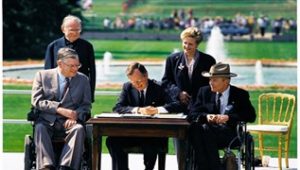 Group photo of signing of Americans with Disabilities Act in 1990. Three individuals are sitting with two of the three in wheelchairs. A man and woman are standing behind them and smiling. Pres Bush is signing the document. Justin Dart is wearing a hat. 
