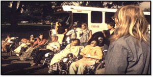 Image of Gang of 19. Men and women in wheelchairs with a bus in the background. 