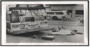 Black and white photo of bus on a street in Colorado. Laying in the street are 4 individuals with disabilities protesting the lack of accessibility on the city buses. 